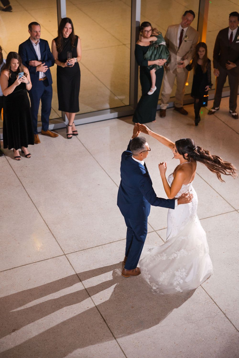 Newlyweds share their first dance while wedding guests look on from the sidelines in elegant formal attire.