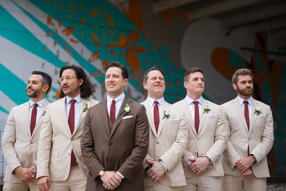 Groomsmen stand in a row wearing tan suits with burgundy ties against a colorful mural wall background.