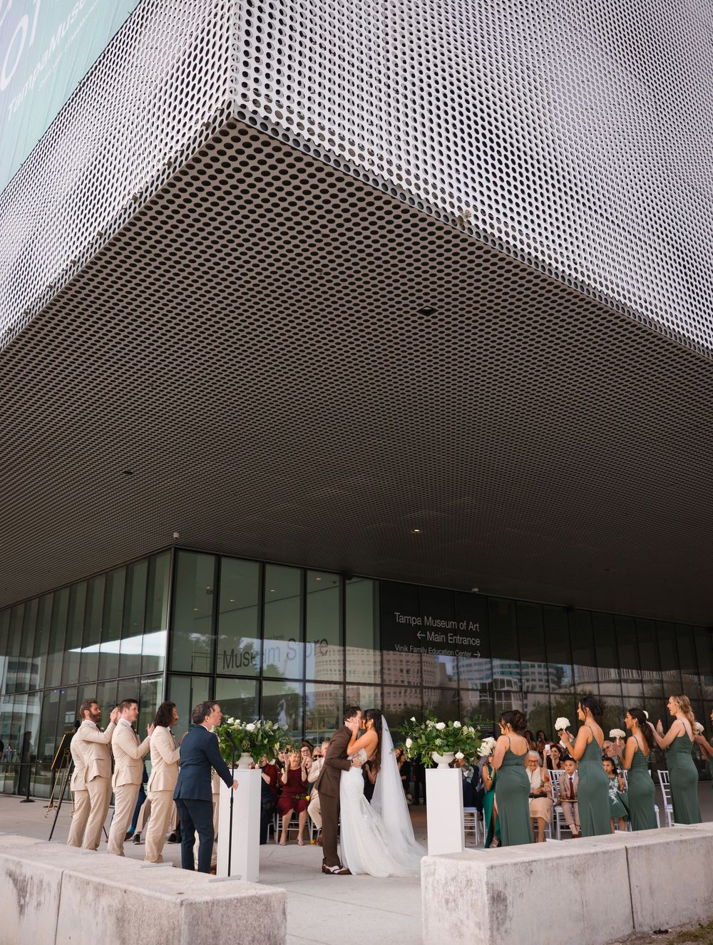 A line of people in formal attire wait outside a modern building entrance with a perforated metal facade.