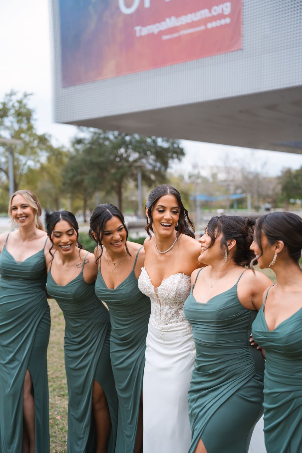 A bridal party wearing matching green dresses poses together outdoors on a sunny day.