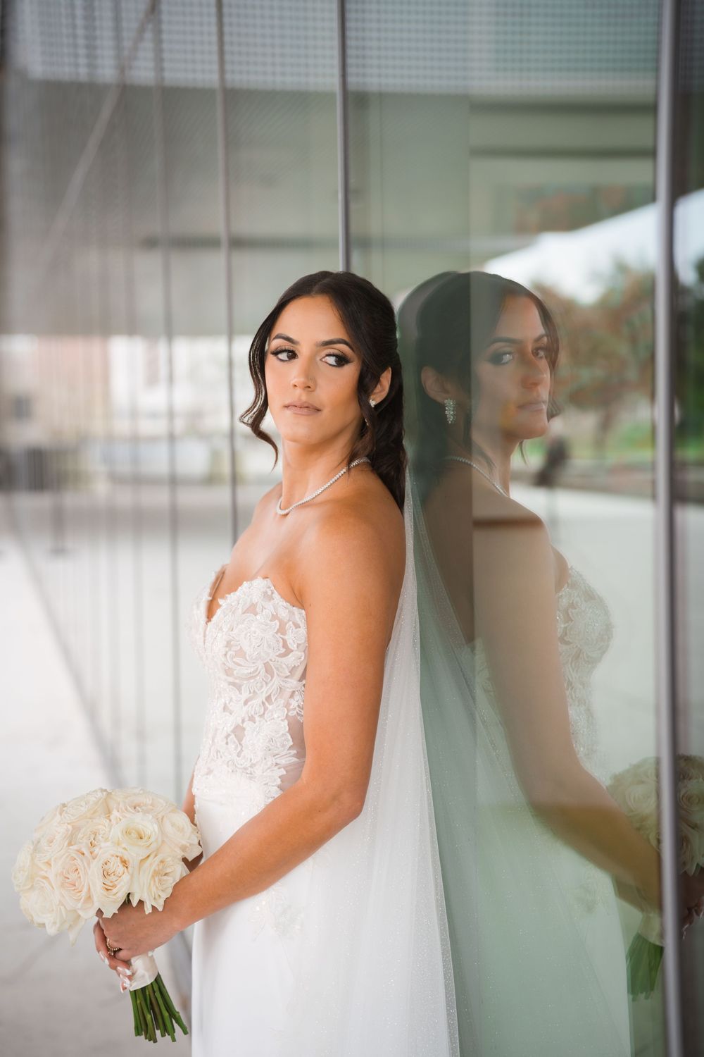 A bride in a beautiful white lace wedding dress poses elegantly by a window holding a bouquet.