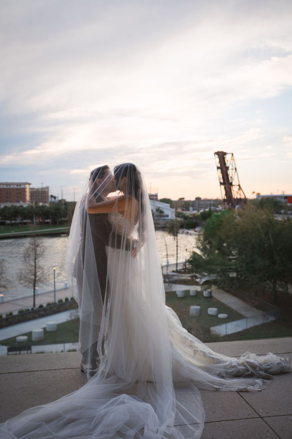 Beautiful wedding scene showing a bride in a flowing white veil overlooking a waterfront cityscape at sunset.