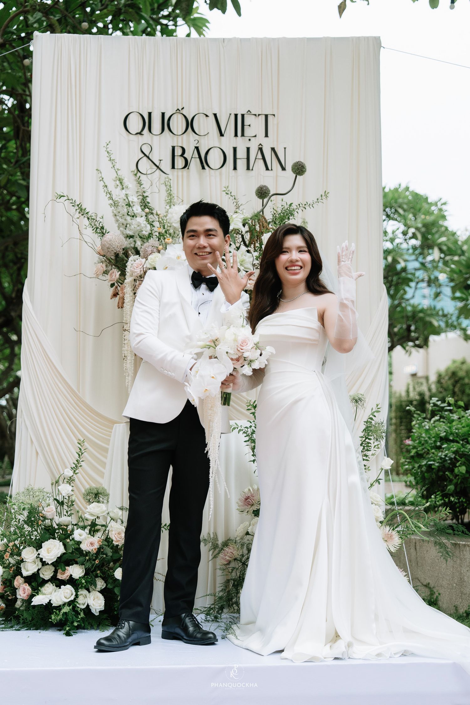 Newlyweds Quoc Viet and Bao Han pose together showing rings at their white floral wedding backdrop outdoors.