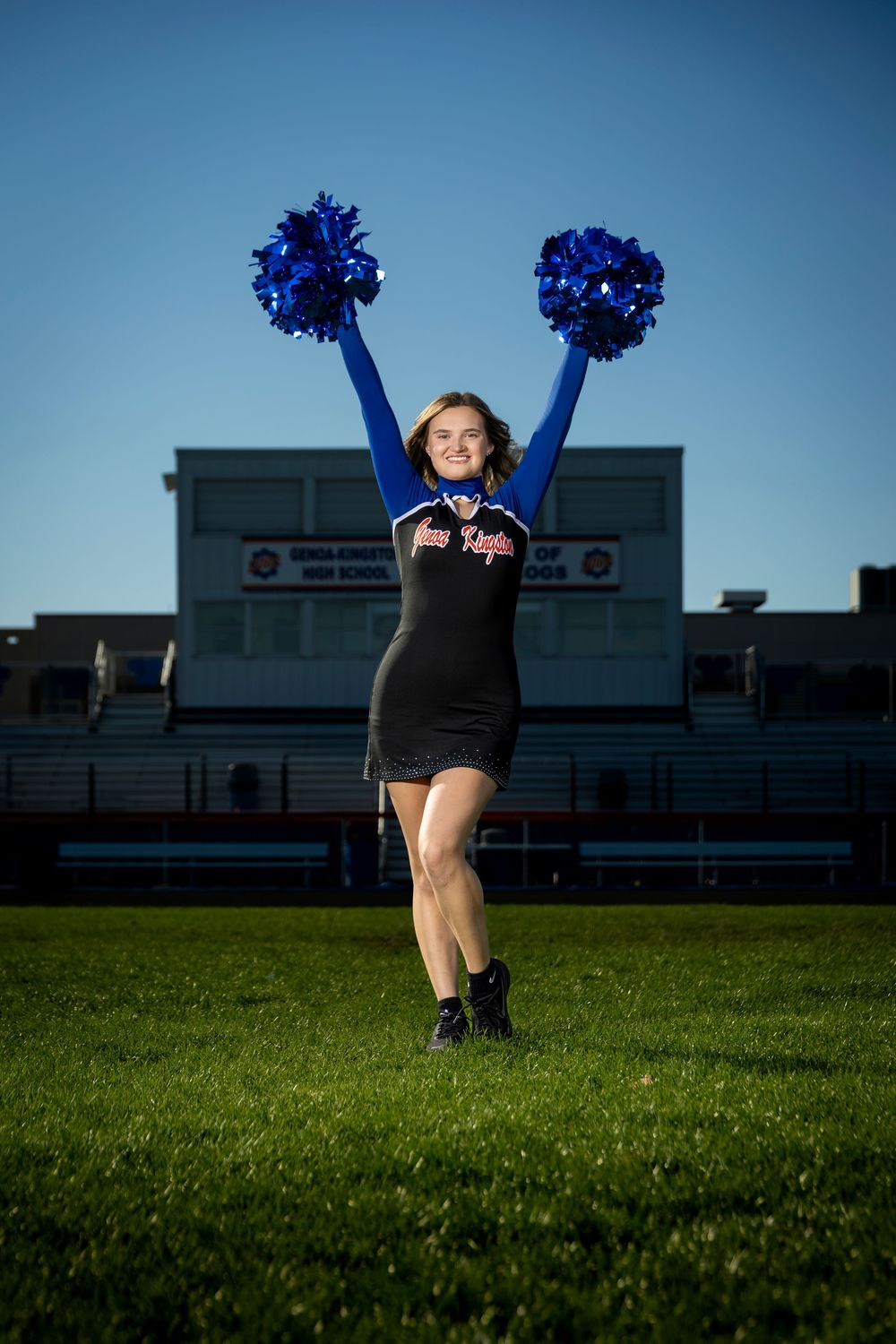 A cheerleader in a black and blue uniform raises blue pom-poms on a green field in front of a high school stadium.