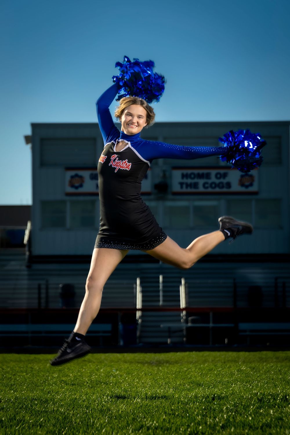 Cheerleader in blue and black uniform jumps mid-air with blue pom-poms at a football stadium at night.