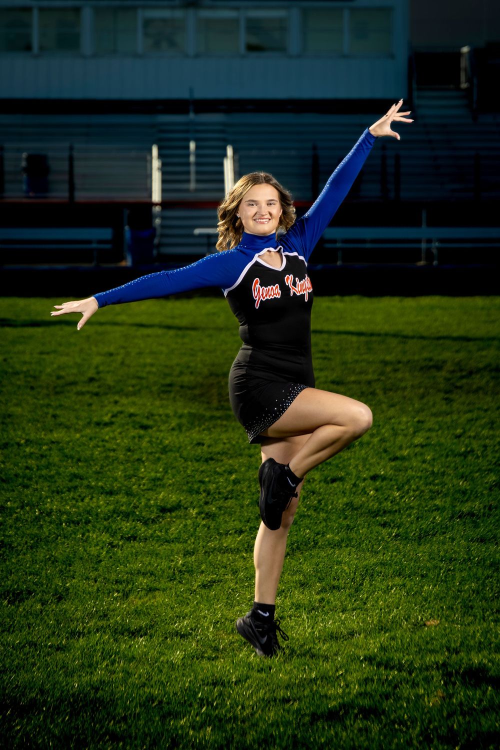 Cheerleader in blue and black uniform poses gracefully on a green field at night, one leg raised with arms extended.