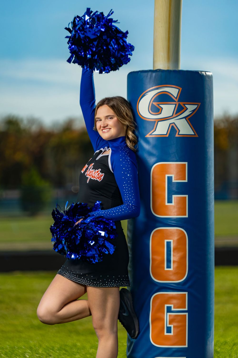 Cheerleader in blue and black uniform poses with pom-poms beside a GK Cougars goal post pad on a football field.