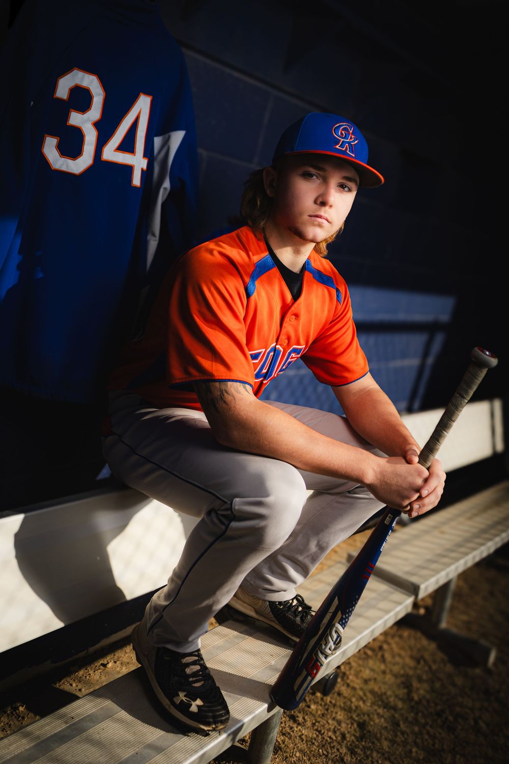 Young baseball player in orange uniform sitting in dugout holding bat, wearing blue cap with team logo.