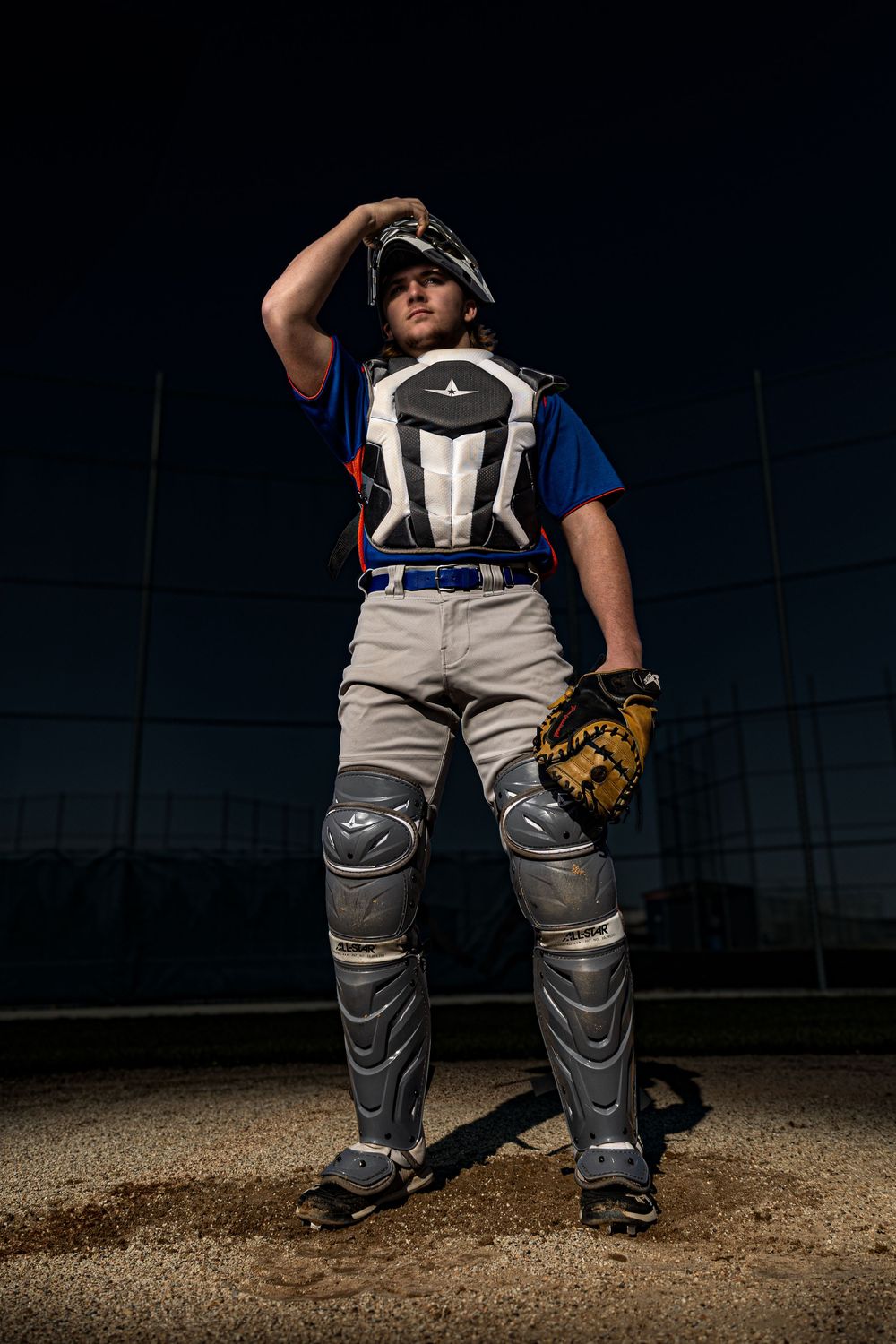 Baseball catcher in full gear poses dramatically at night, wearing chest protector, leg guards, and mitt.