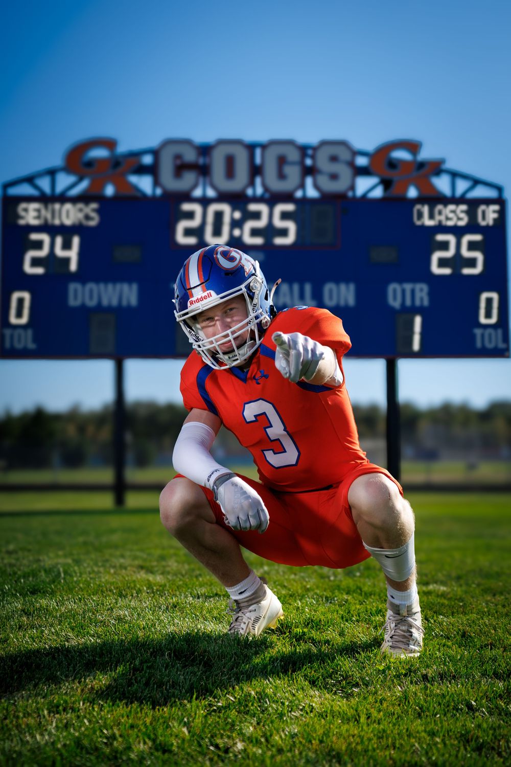 Football player in orange jersey crouches on grass field with scoreboard in background.