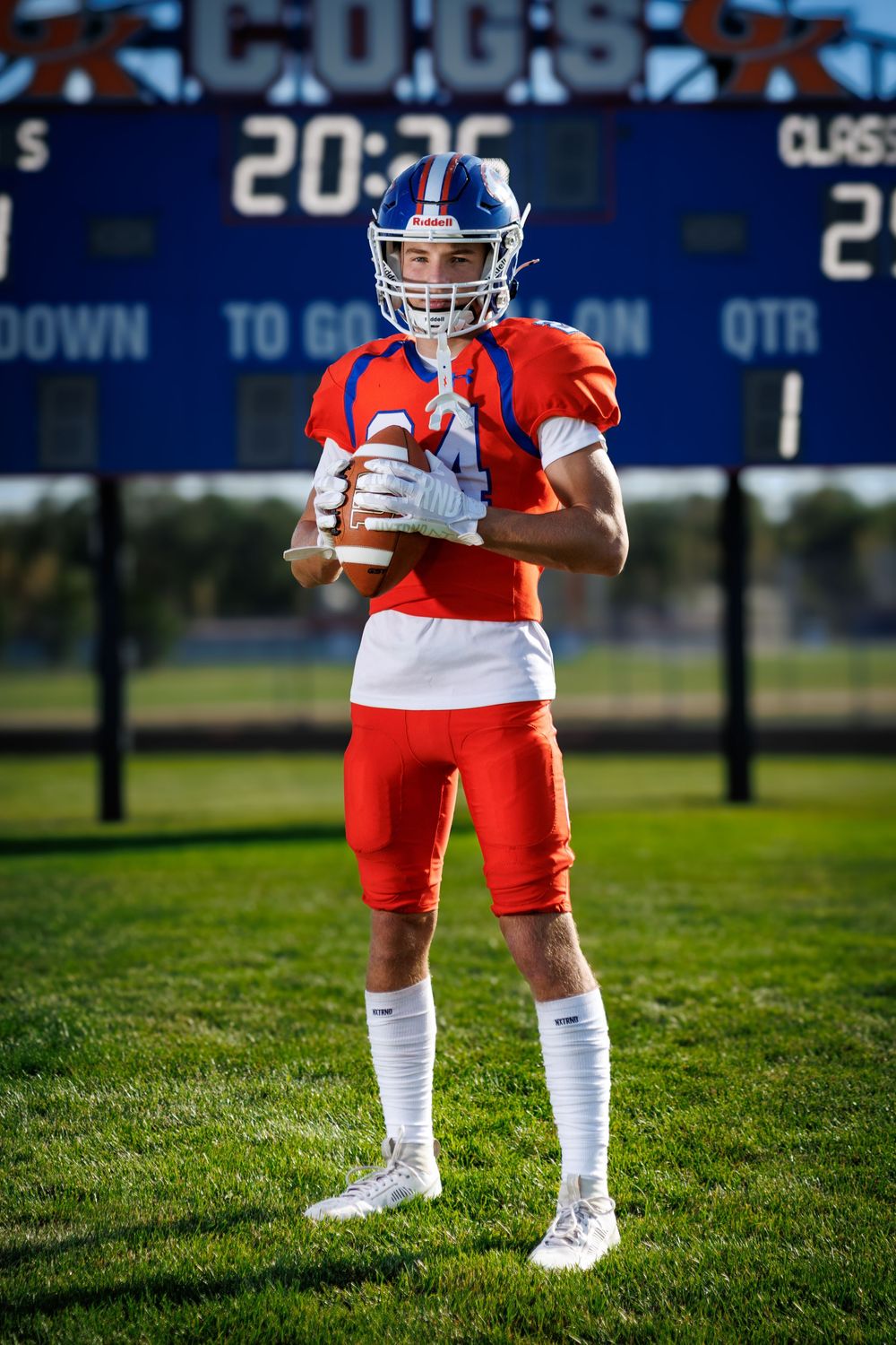 Football player's legs in orange uniform and white socks on green field with scoreboard backdrop.