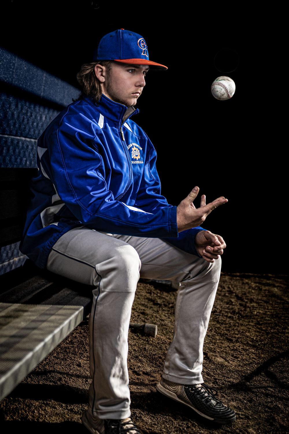 Baseball player in blue uniform tosses ball while sitting in dugout shadows.