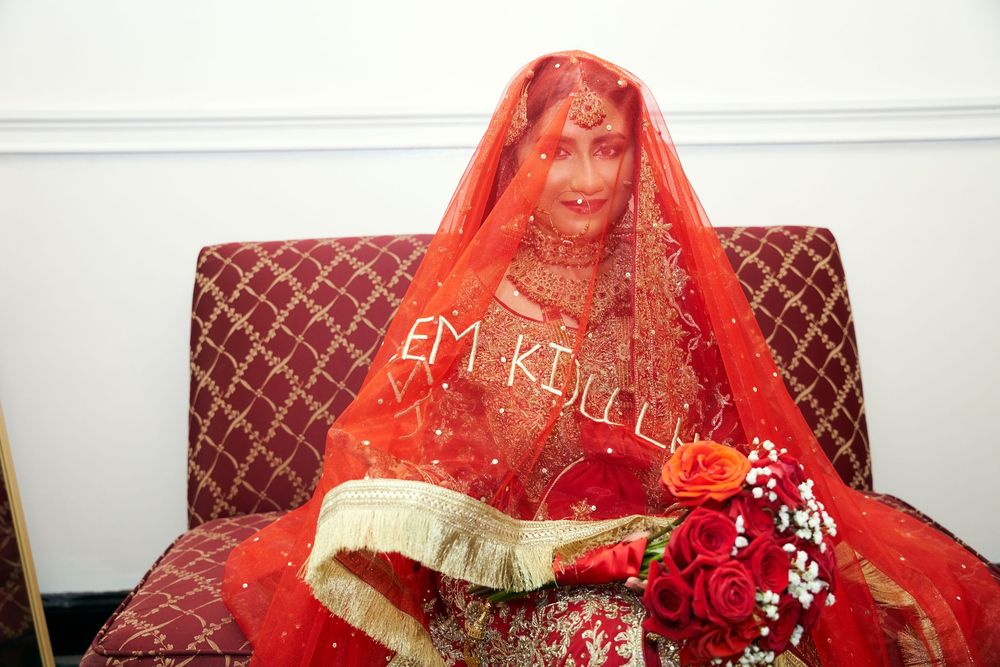 Traditional South Asian bride wearing a vibrant red and gold bridal outfit with matching veil seated on brown chair. at Antuns by Minar
