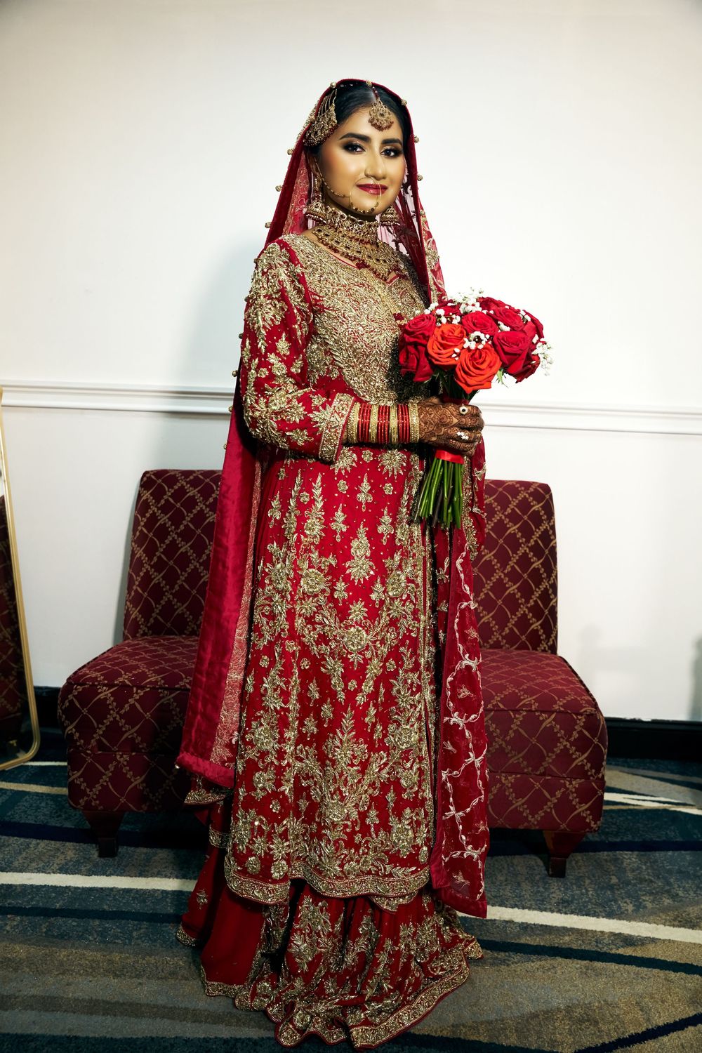 Traditional South Asian bride in ornate red and gold embroidered lehenga holds vibrant red bouquet against quilted bench at Antuns by Minar