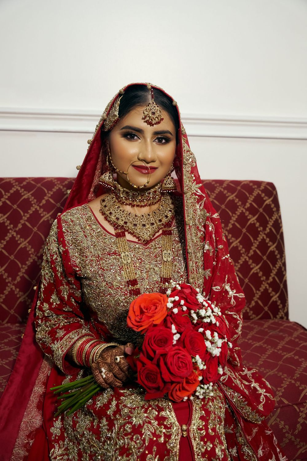 Traditional Indian bridal attire in red and gold with embellished lehenga, jewelry and bouquet of red roses. at Antuns by Minar