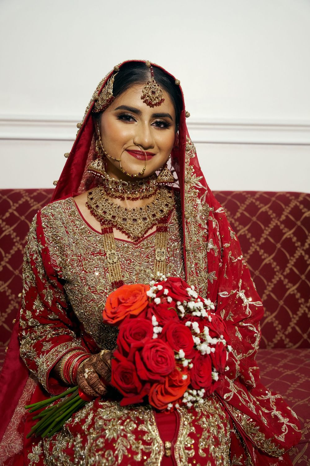 Traditional Indian bride in red and gold lehenga holds vibrant red roses and baby's breath bouquet. at Antuns by Minar