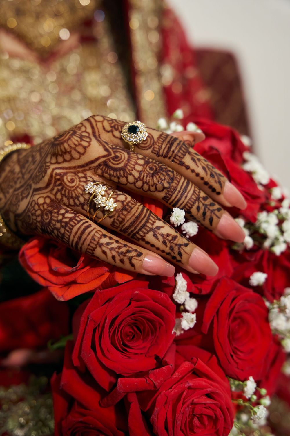 Ornate henna-decorated hands with diamond rings resting on vibrant red roses and pearl jewelry. at Antuns by Minar