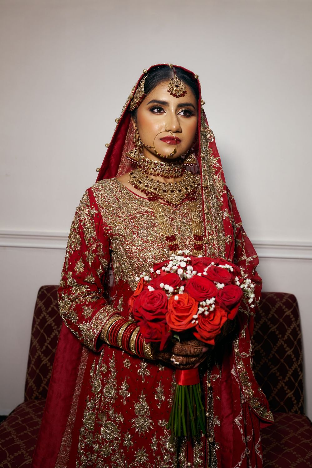 A bride in a traditional red and gold South Asian wedding lehenga holds a vibrant bouquet of red roses and baby's breath. at Antuns by Minar