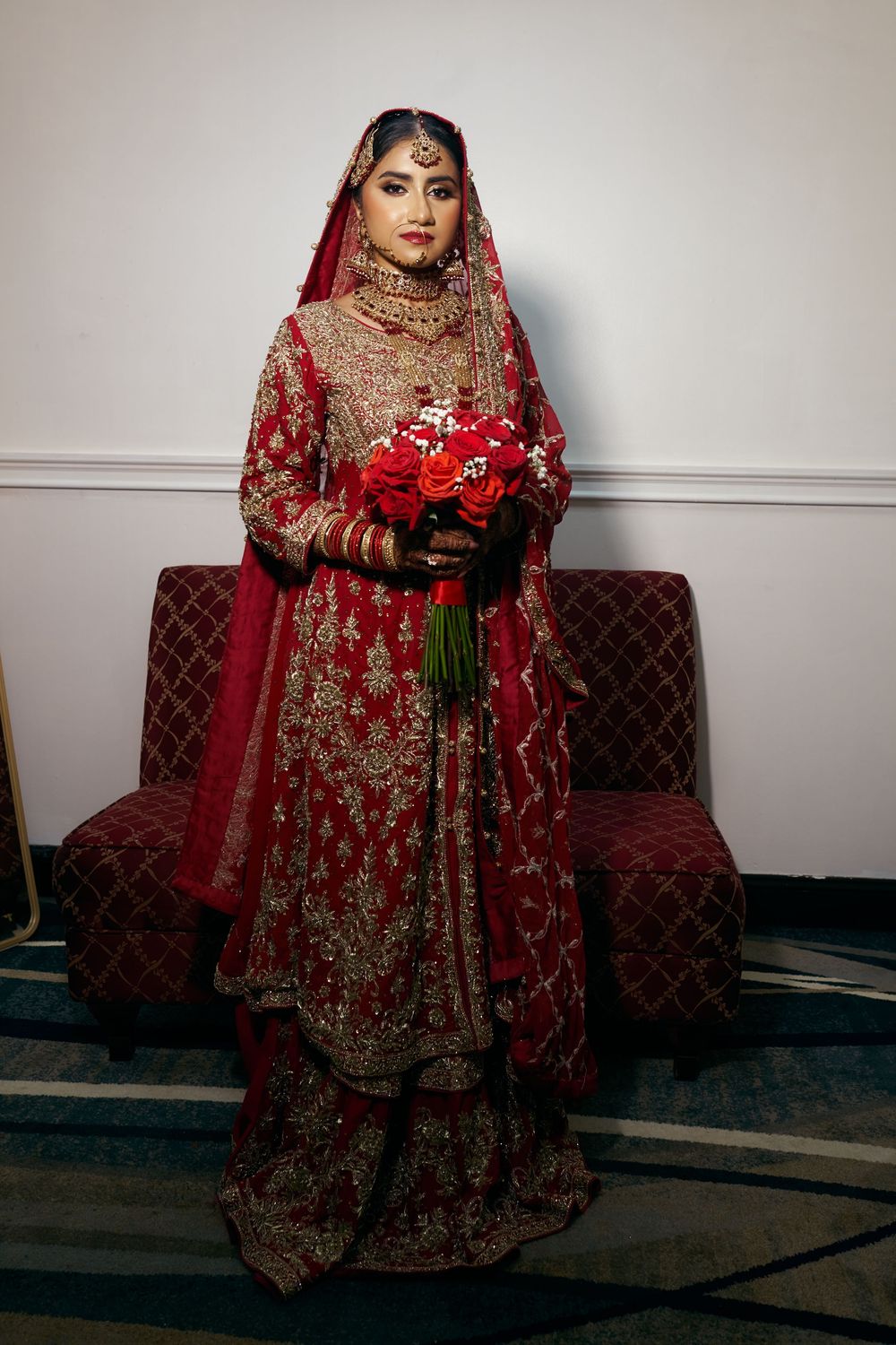 Traditional South Asian bride in ornate red and gold embroidered lehenga holds bouquet while standing against dark couch. at Antuns by Minar