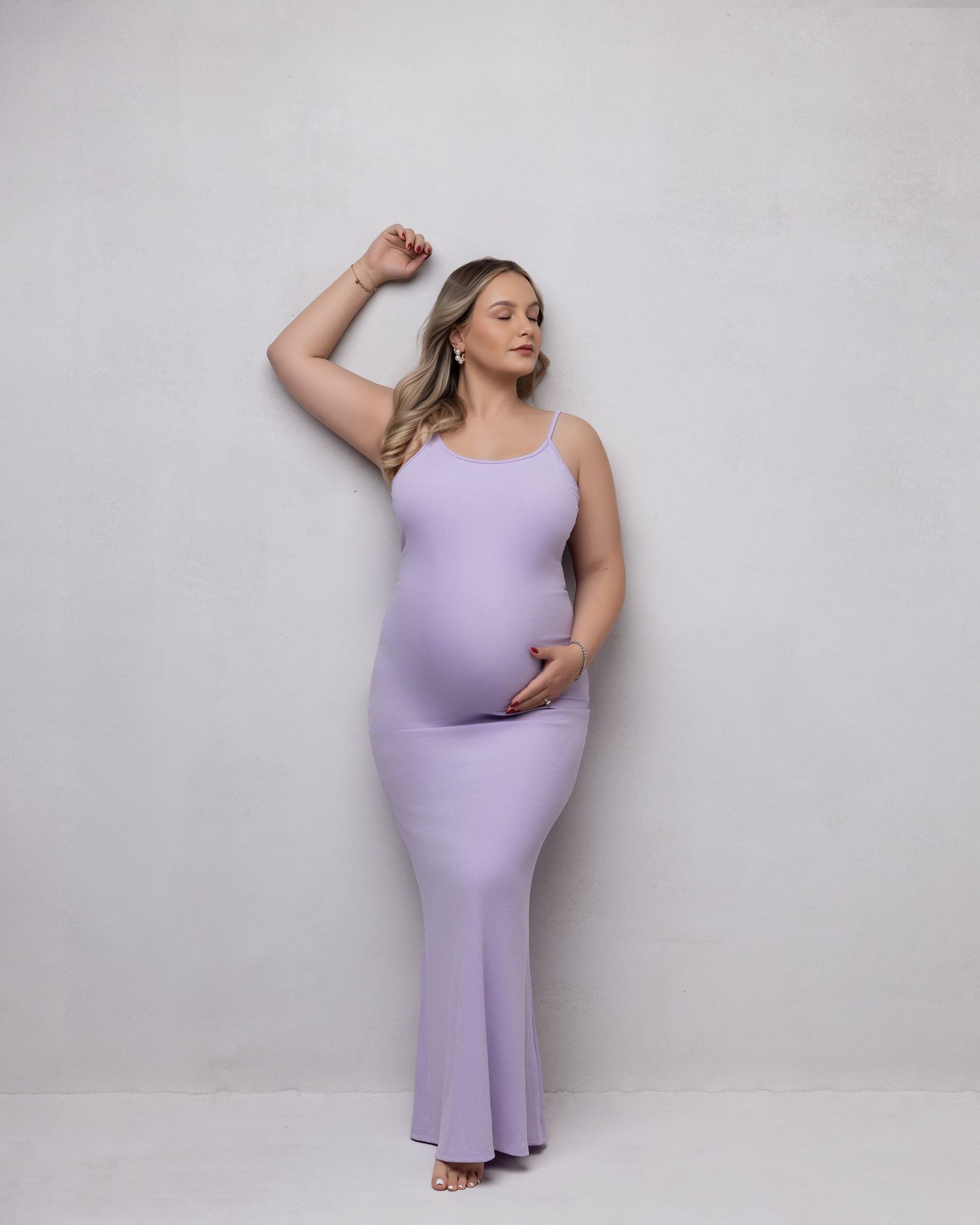 A person wearing a fitted lavender maxi dress poses against a light gray background while expecting.