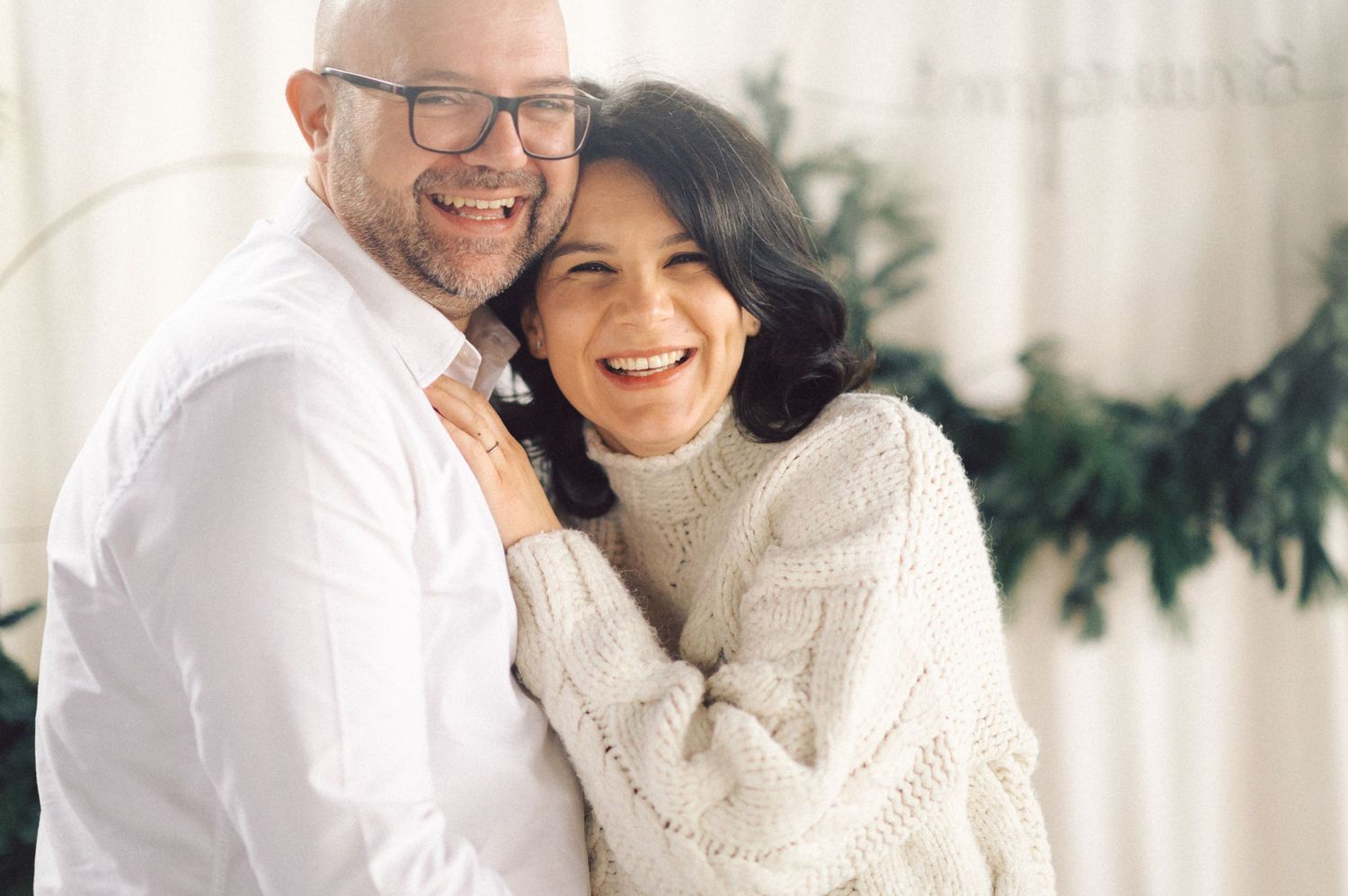 Happy couple in white clothes sharing an intimate moment with holiday greenery and candles in background.