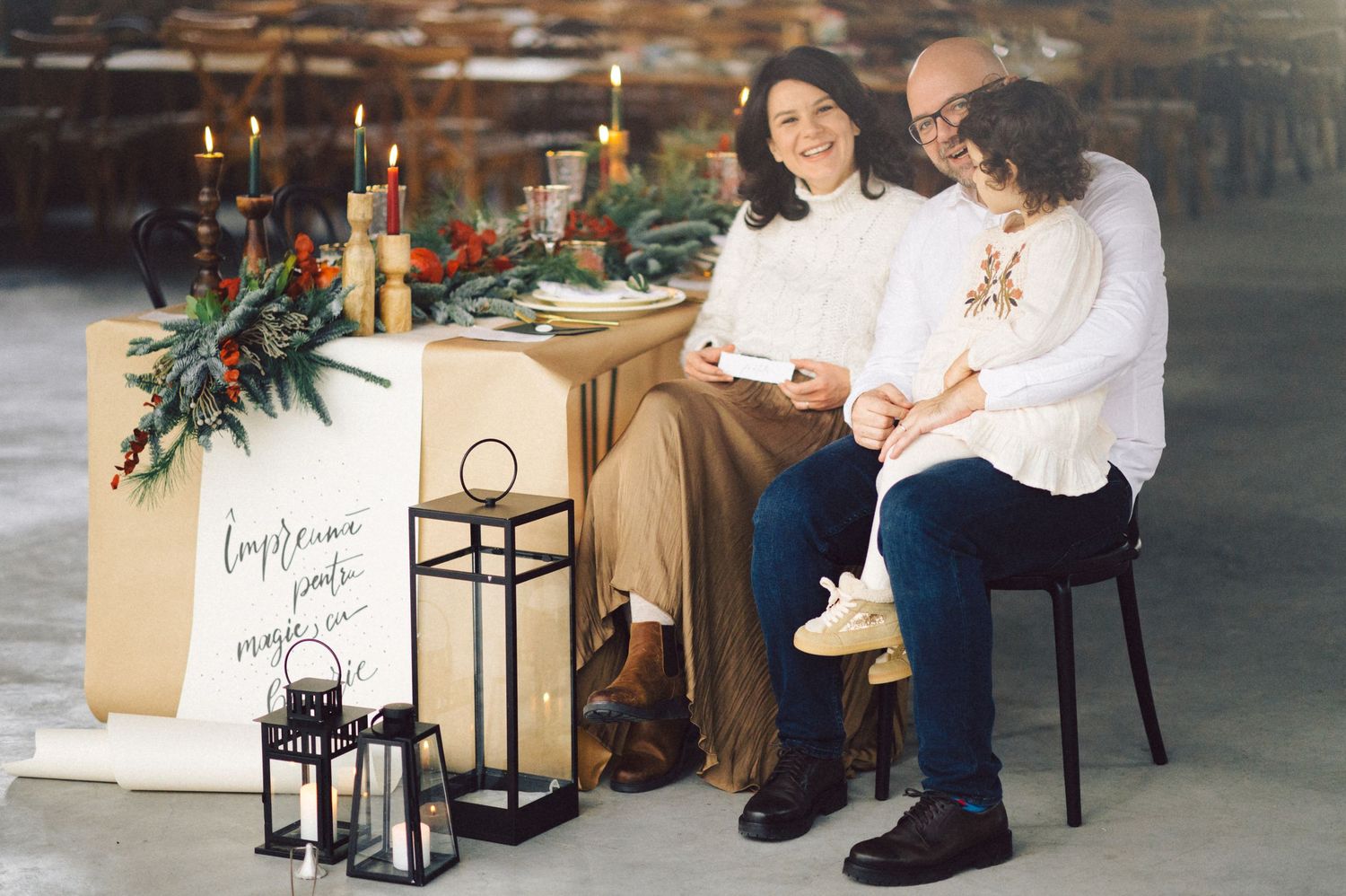 Two people in white sweaters sit at a decorated table with candles, greenery and lanterns for a cozy holiday celebration.