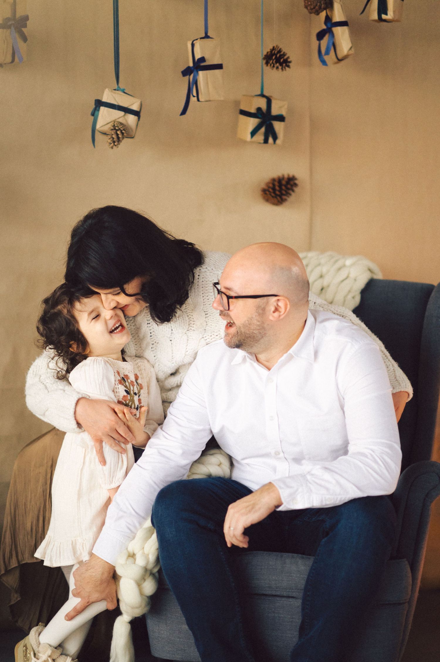 A couple shares a tender moment on a couch under hanging holiday decorations and pinecones in a warm setting.