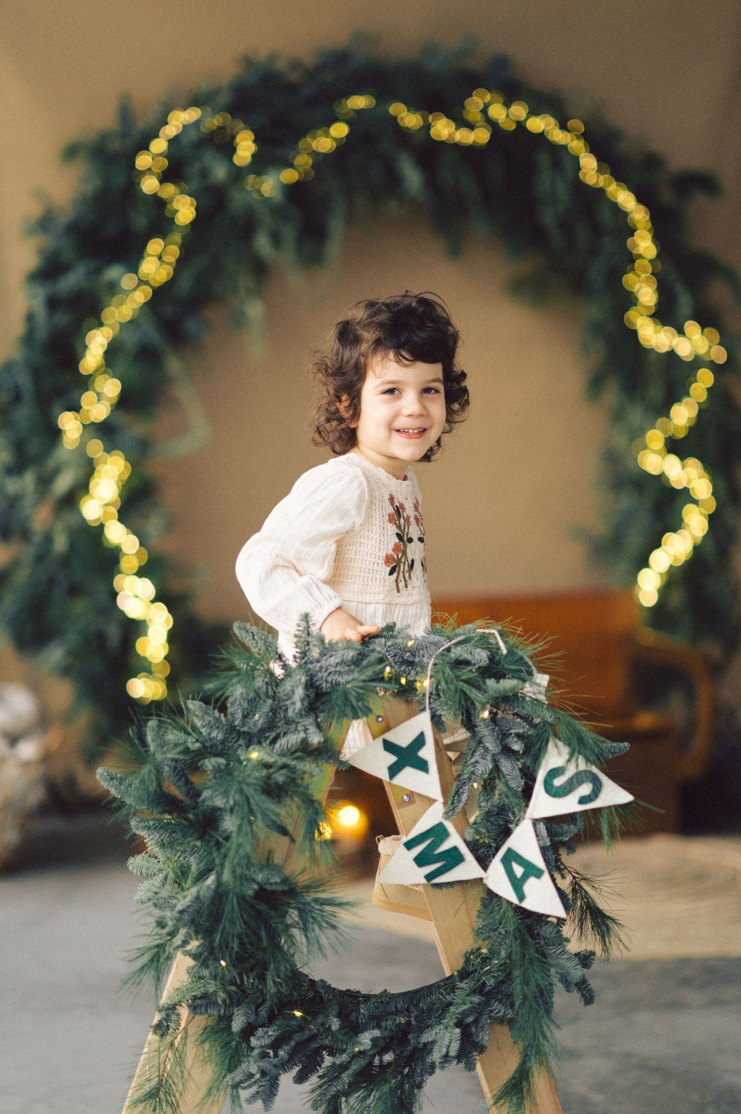 Holiday portrait with evergreen wreath arch and decorative Christmas lights in a warm indoor setting.