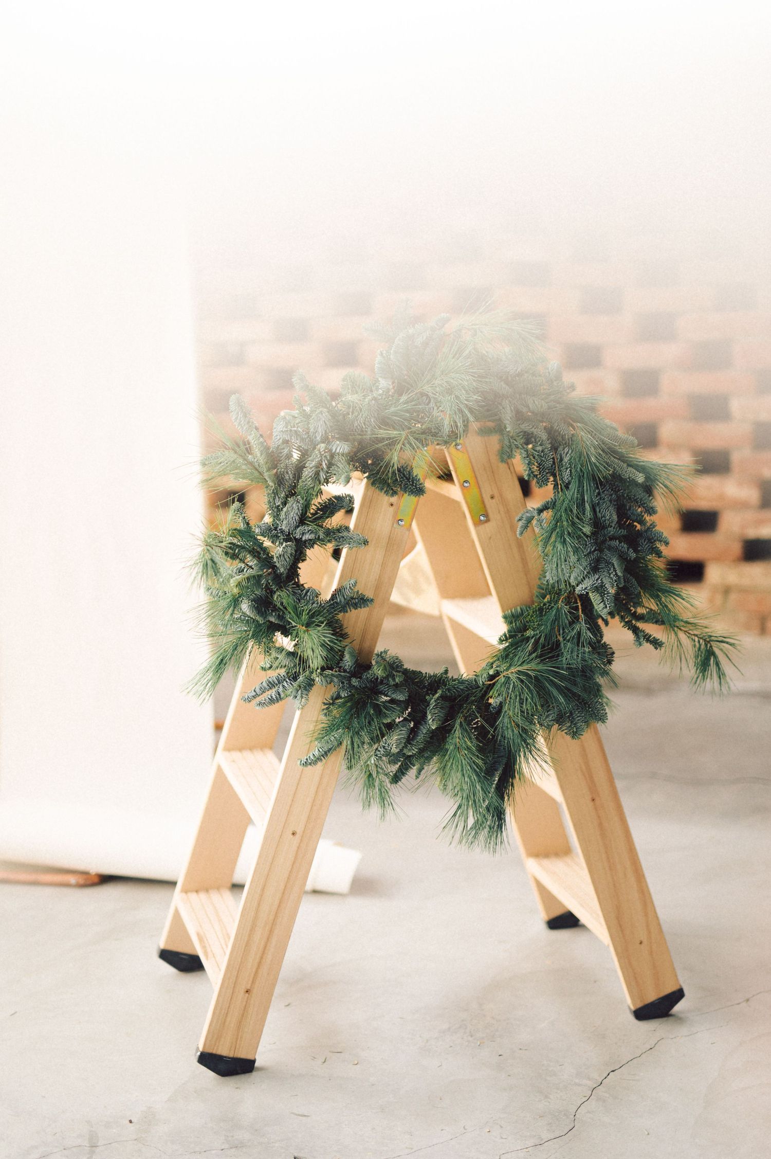 A rustic holiday wreath made of fresh pine branches rests on top of a wooden stepladder against a brick wall.