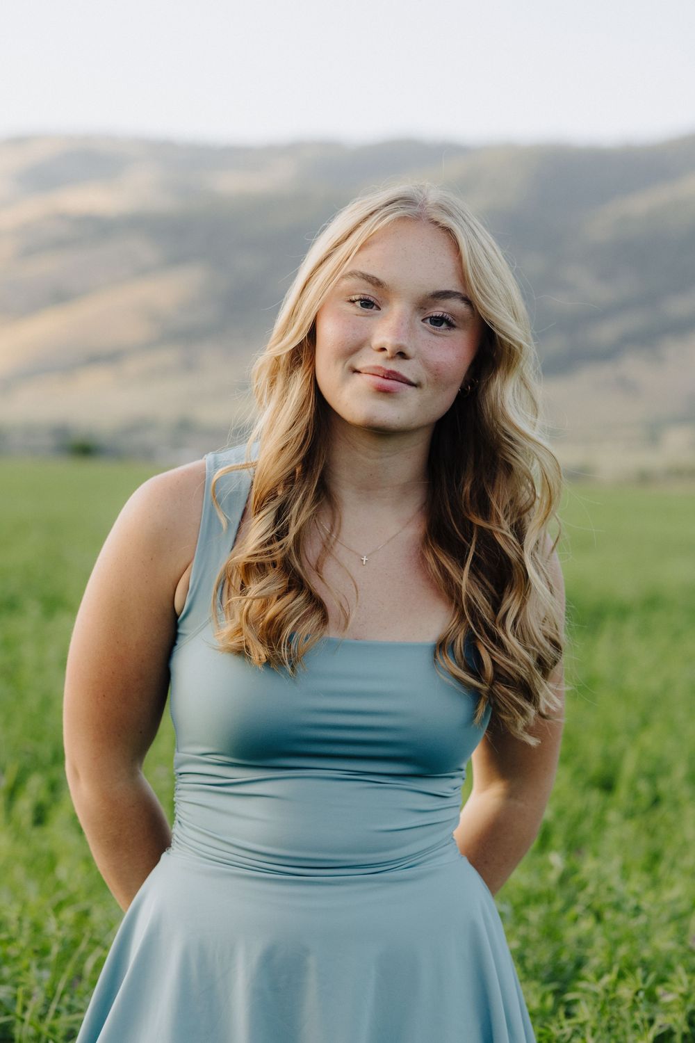 A person in a light blue dress poses in a green field with mountains in the background.