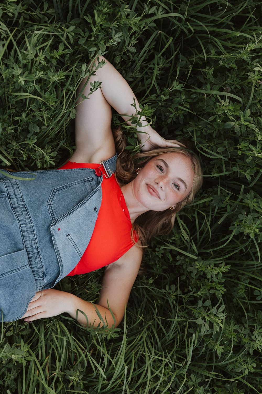 Person in red top and denim lying in tall green grass during a summer photoshoot.