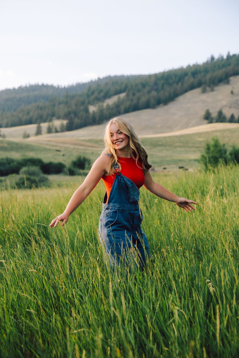 Playful summer portrait in tall grass wearing overalls against mountain backdrop.