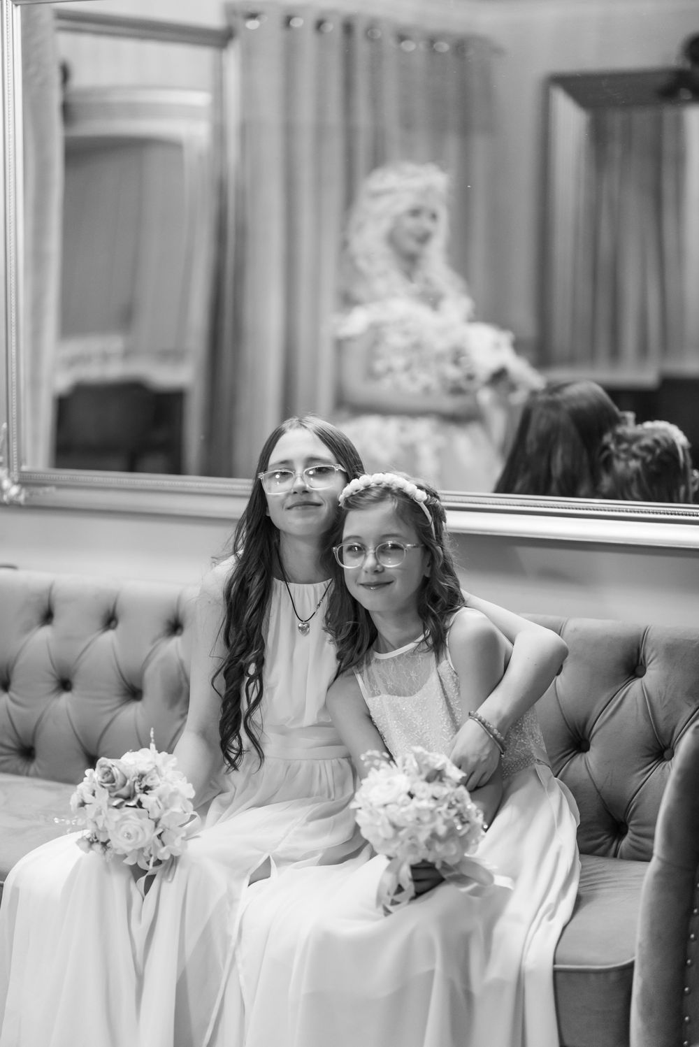 Young flower girls in white dresses sit together on an elegant tufted couch during a wedding celebration.