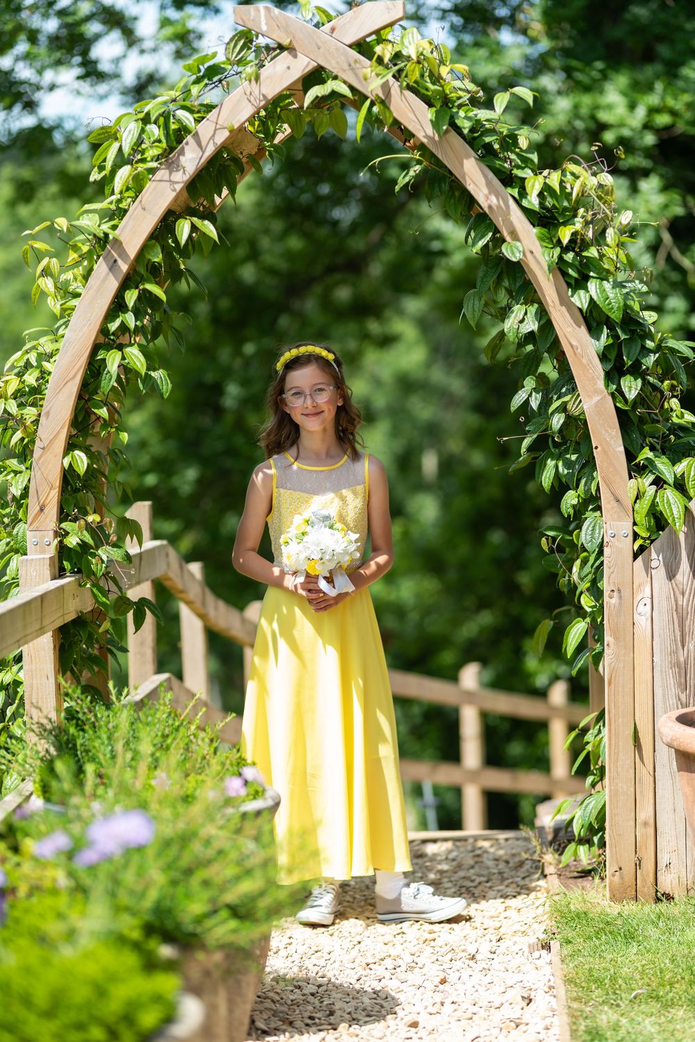 Yellow-dressed attendants stand under wooden garden arch decorated with greenery.