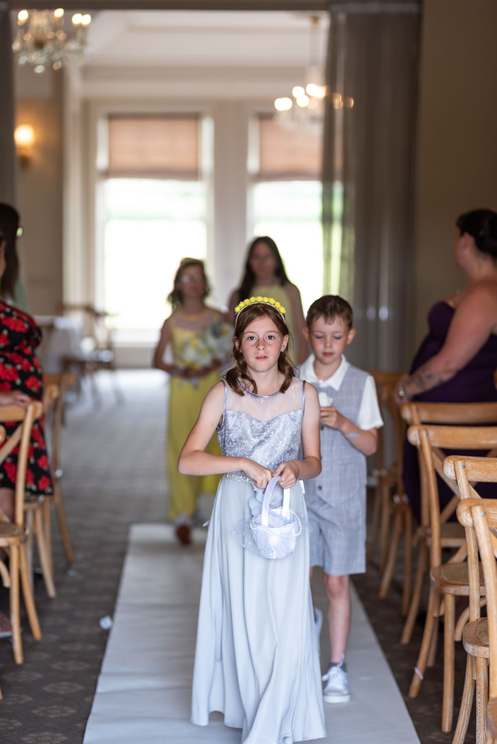 Young attendants in light colored dresses walking down aisle at indoor ceremony.