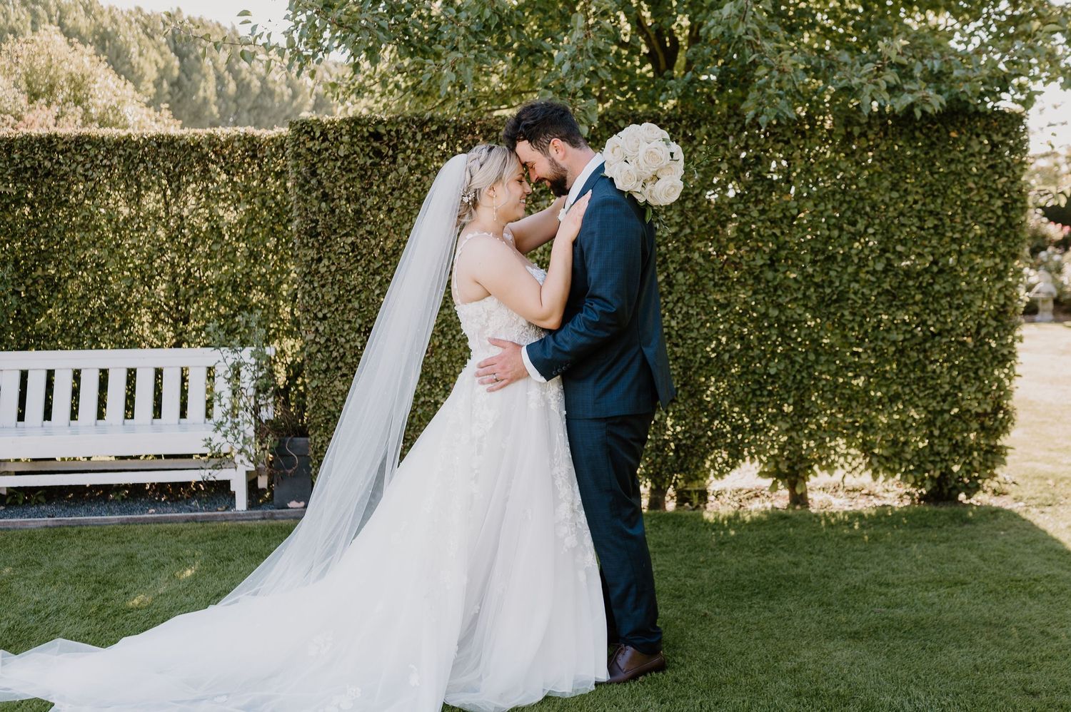 A romantic wedding couple embraces in front of a lush garden hedge with a white bench nearby.