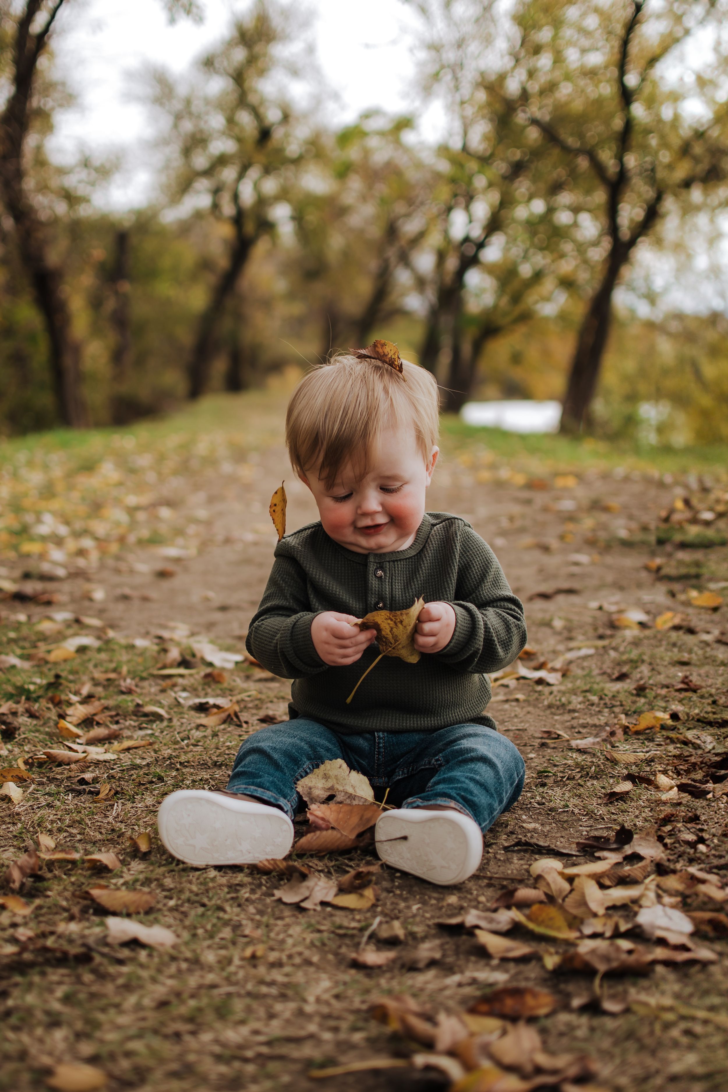 Little one sitting on leaf-covered ground in green sweater and jeans during autumn exploring nature.