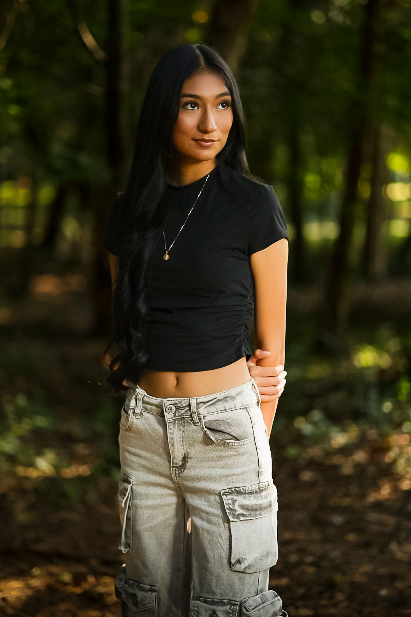A person poses in a black crop top and light cargo pants outdoors in a wooded setting at Gulley Park
