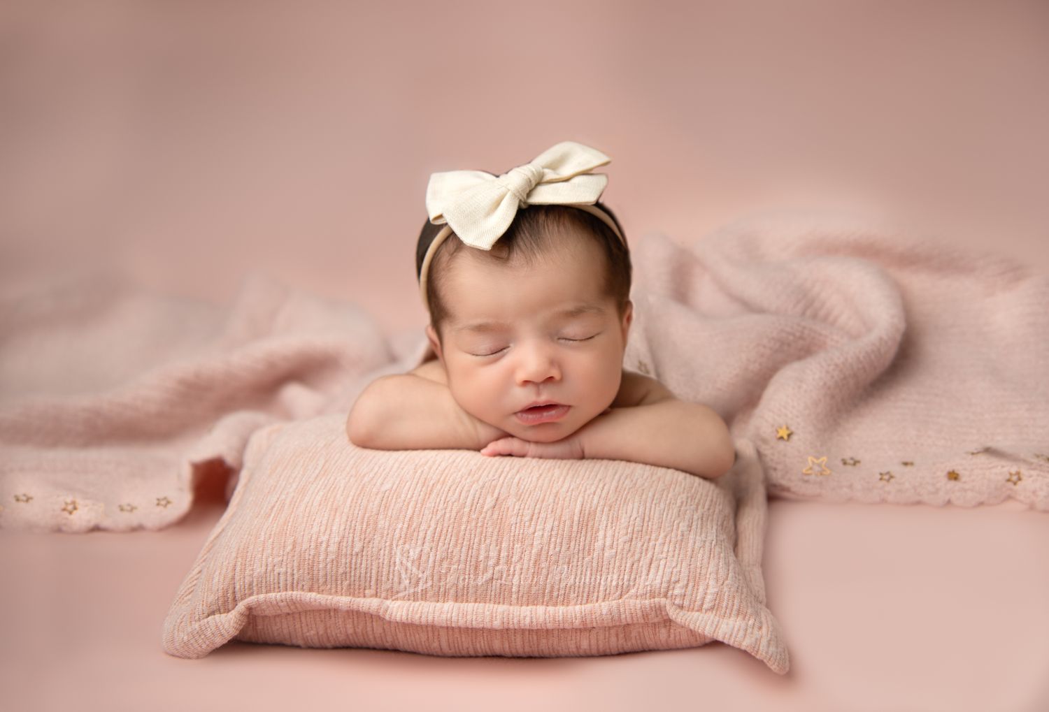 A sleeping newborn baby girl with an off white headband wrapped in soft pink fabric lies peacefully on a textured blanket.