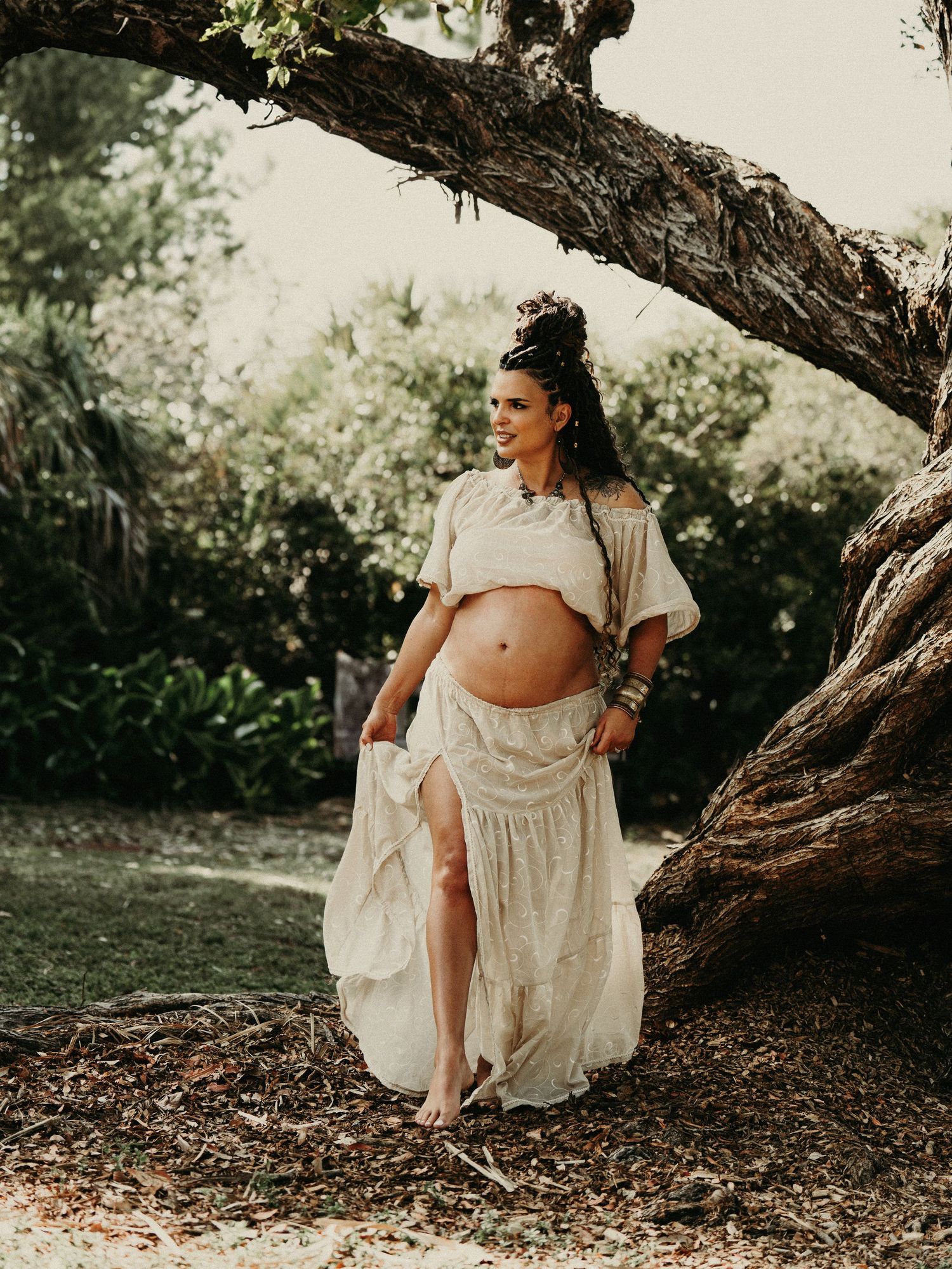 An expectant woman in a flowing white dress poses gracefully beside a large tree trunk.