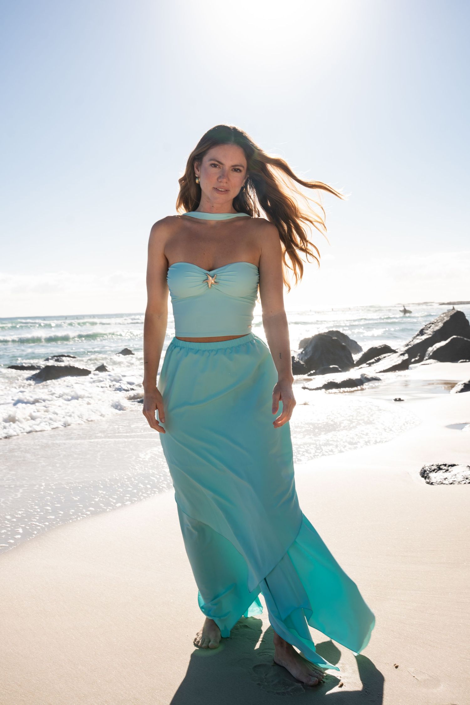 A swimwear model in a flowing turquoise dress poses on a beach lifestyle shoot with rocky outcrops and a Gold Coast beach in the background.
