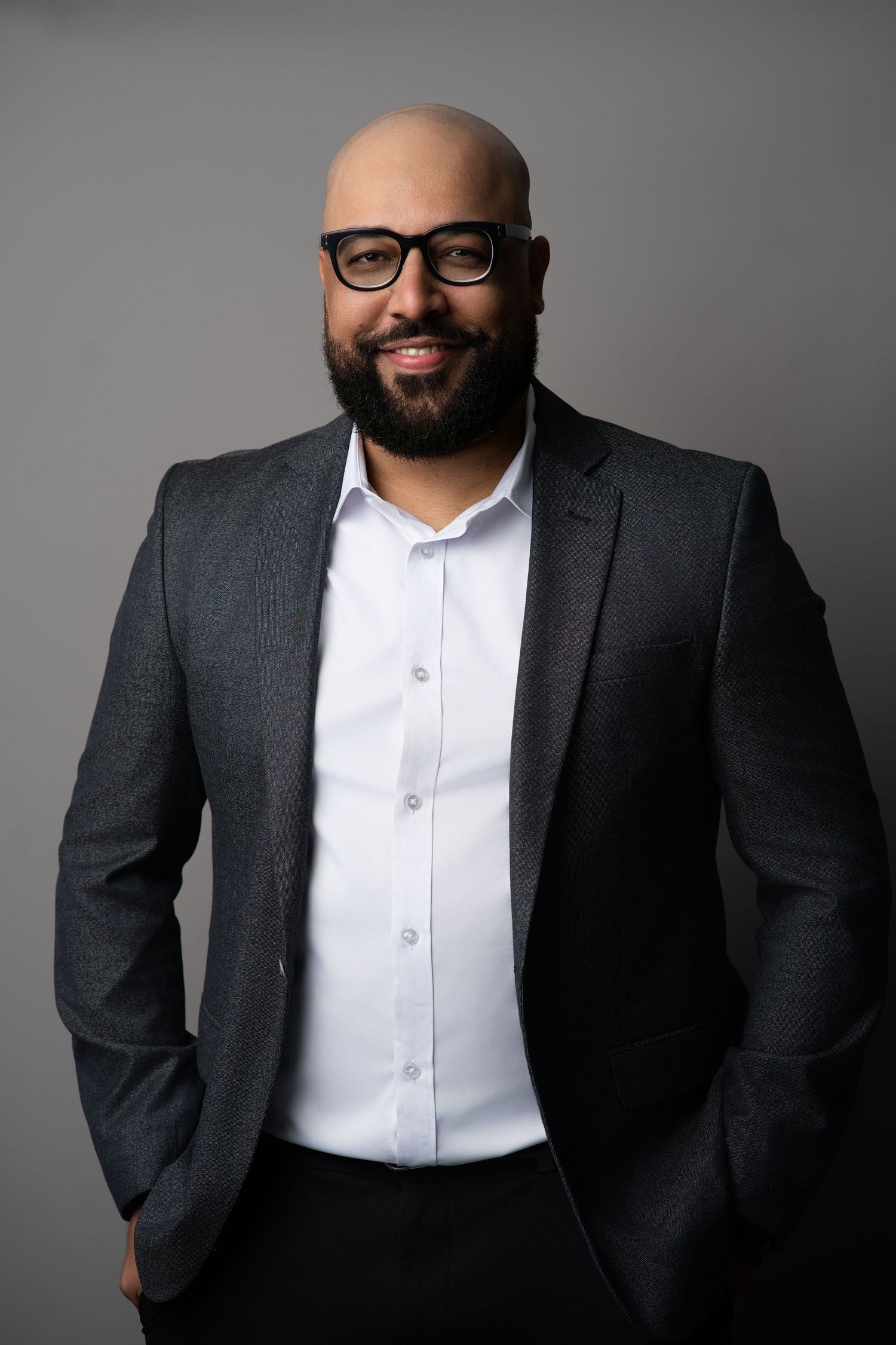Professional headshot of someone in dark suit and glasses with warm expression against gray background.
