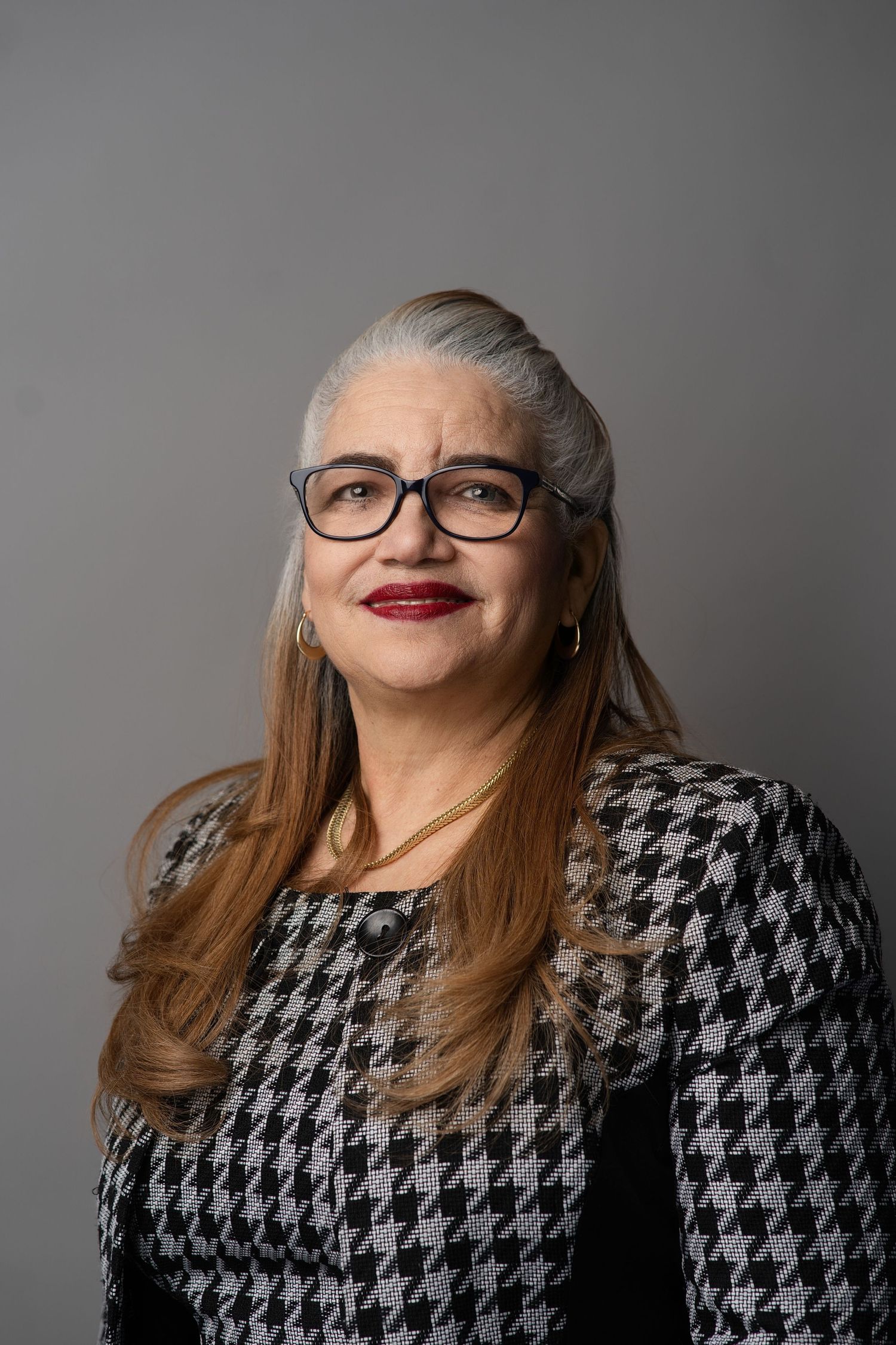 Professional portrait featuring a houndstooth pattern jacket, glasses, and sophisticated styling against a gray background.