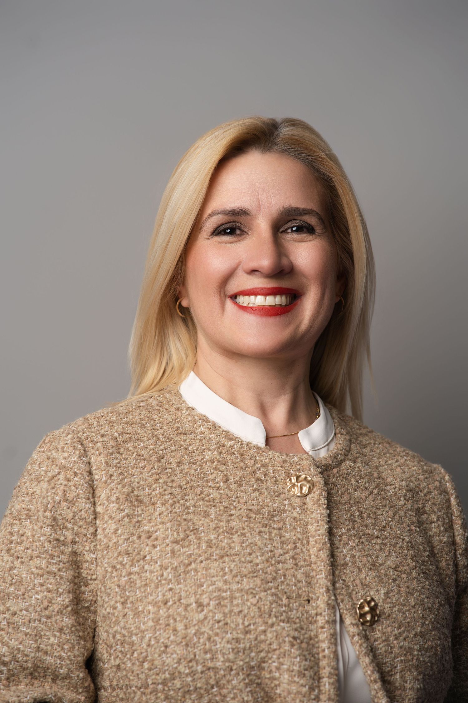 Professional portrait showing beige textured jacket, white collar, and bright smile against neutral background.