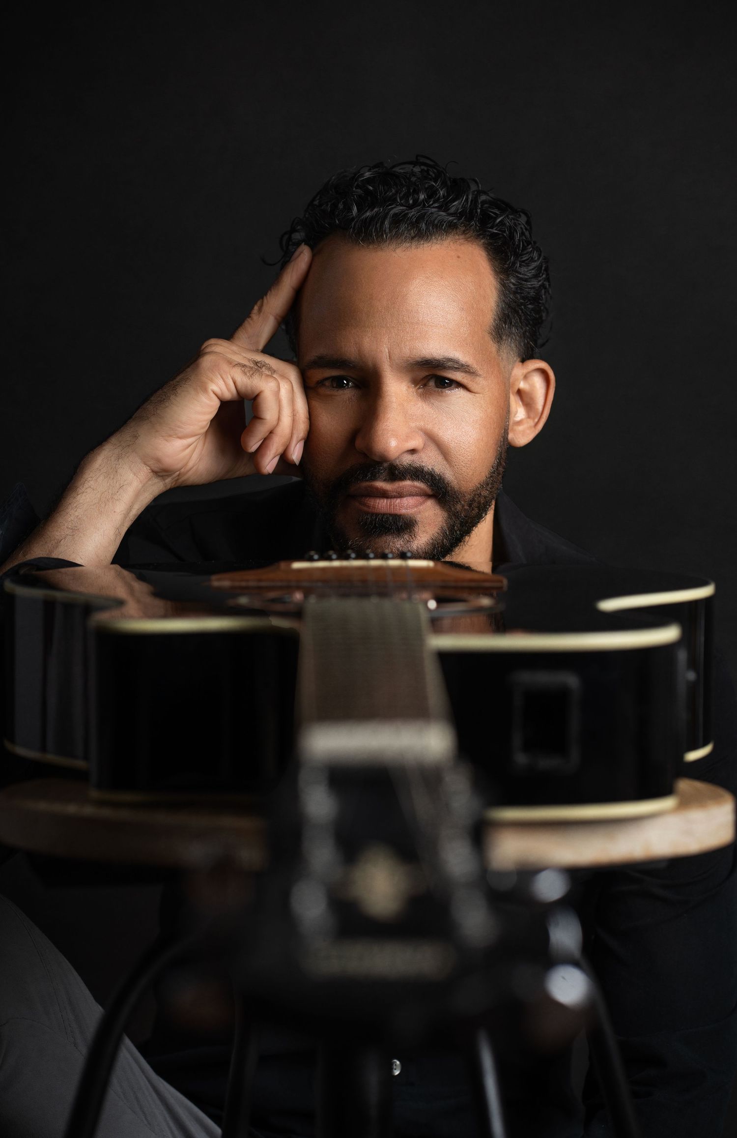 Artistic portrait with guitar in foreground showing contemplative pose against dark background.