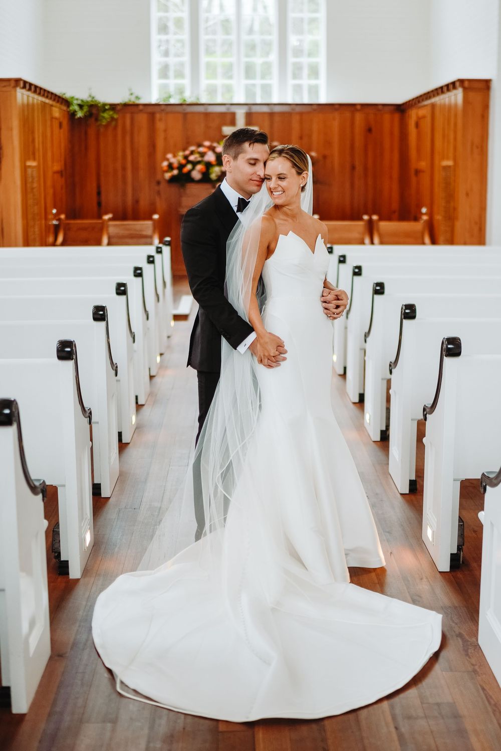 A wedding couple poses elegantly in a church aisle with wooden paneling and white pews.