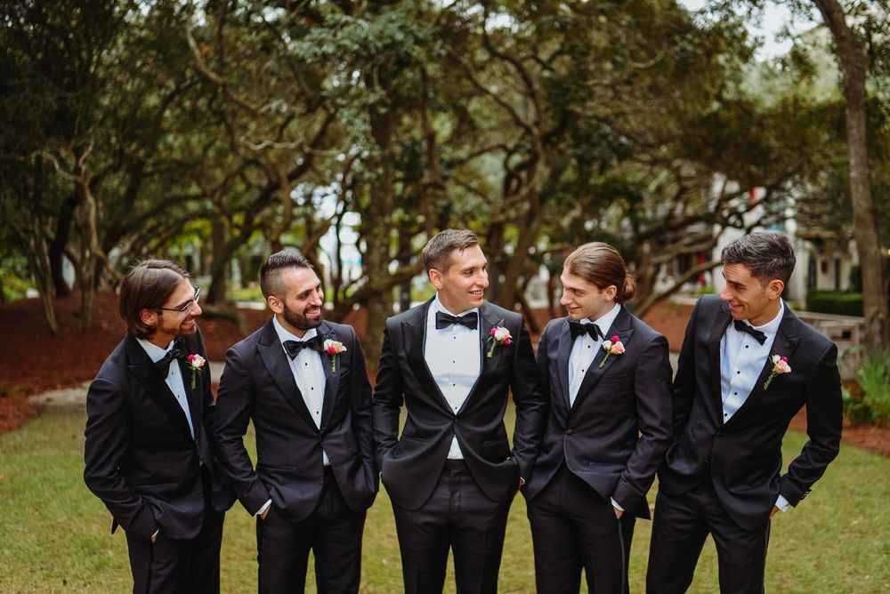 Groomsmen in black tuxedos stand together laughing and smiling outdoors in a park setting.