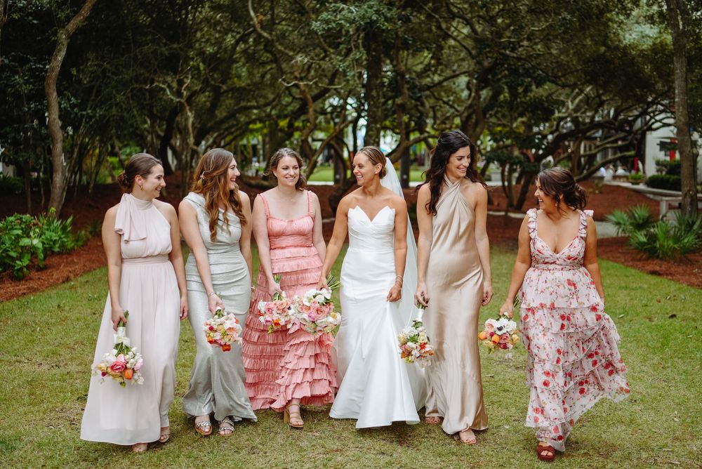 Group of bridesmaids wearing white and floral dresses pose together on grass holding colorful bouquets.