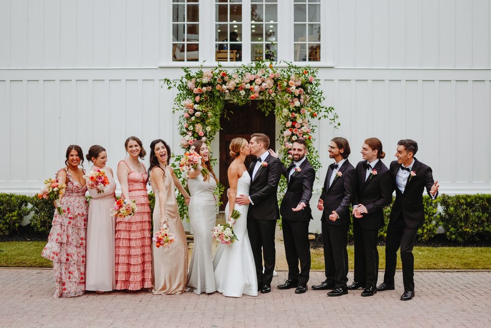 Wedding party poses in front of white church with bridesmaids in coral dresses and groomsmen in black suits.