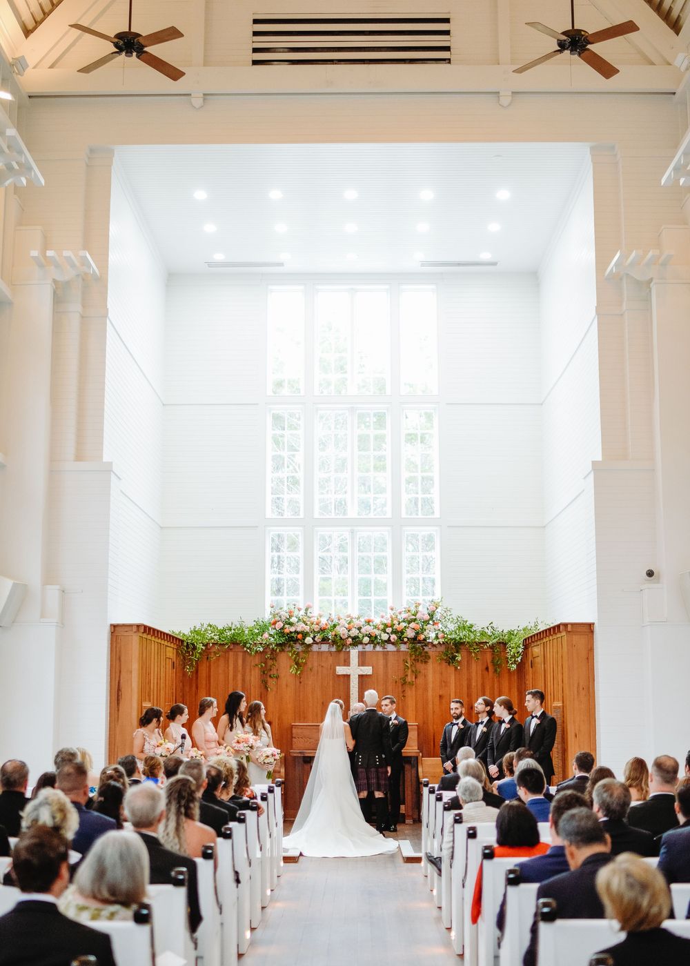 A wedding ceremony takes place in a bright chapel with high ceilings and natural light streaming through windows.
